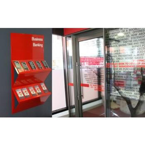 A red freestanding wall-mounted display kiosk with multiple small product slots and a sign reading 'Business Banking,' shown beside glass doors in an indoor setting.